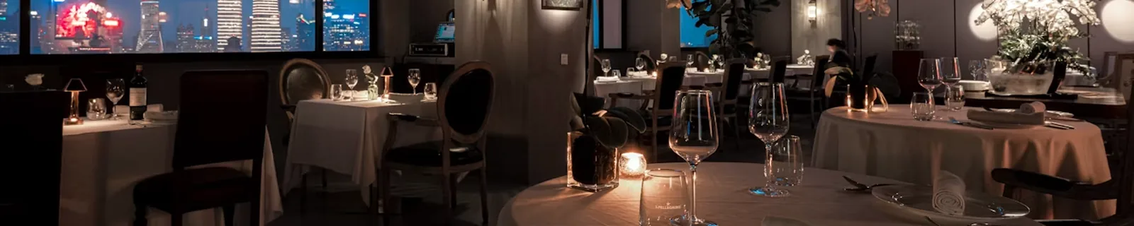 A fine-dining room at night with set tables and a city skyline through the windows